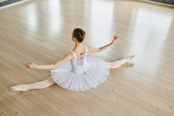 Boven hoek van de rug van sierlijke ballerina in witte tutu strekken haar armen en benen terwijl zitten op de vloer van ruime klaslokaal - Foto, afbeelding