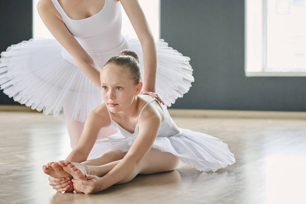 Söpö nuorekas tyttö tekee vaivaa, kun taivutus eteenpäin yli venytetty jalat, kun taas nuori baletti ohjaaja auttaa häntä harjoituksen aikana - Valokuva, kuva