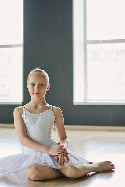 Menina muito jovem em tutu branco e meias bege sentado no chão de sala de aula espaçosa ou estúdio de dança e olhando para a câmera - Foto, Imagem