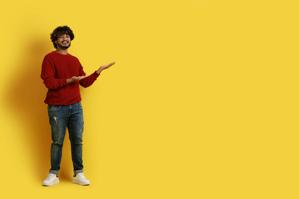 Nice offer. Smiling handsome young indian guy with curly hair in casual outfit hipster posing over yellow studio background, pointing at copy space for advertisement, full length - Photo, Image