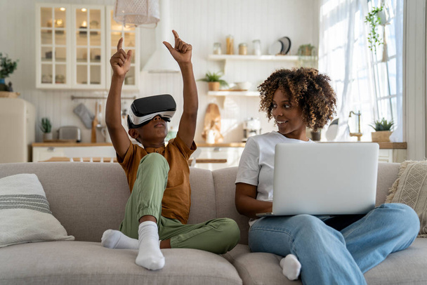 Современная цифровая семья. Счастливая афроамериканская семья мать и сын отдыхают на диване с гаджетами, ребенок играет в виртуальной реальности, сидя рядом мама работает на ноутбуке. Концепция VR и детей - Фото, изображение