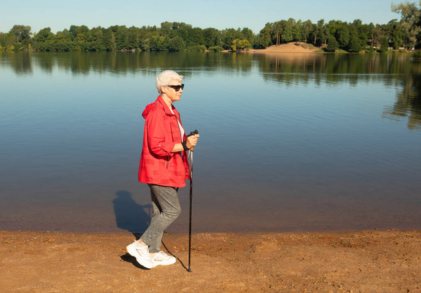 Grey-haired woman wearing red jacket walking with tracking sticks on the beach near lake, summer day - Fotó, kép