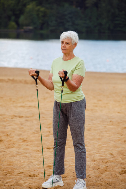 Old woman wear sportswear doing exercises outdoor on the beach in the morning using resistance rubber bands. Healthy lifestyle, active retired life and sporty time. - Fotoğraf, Görsel