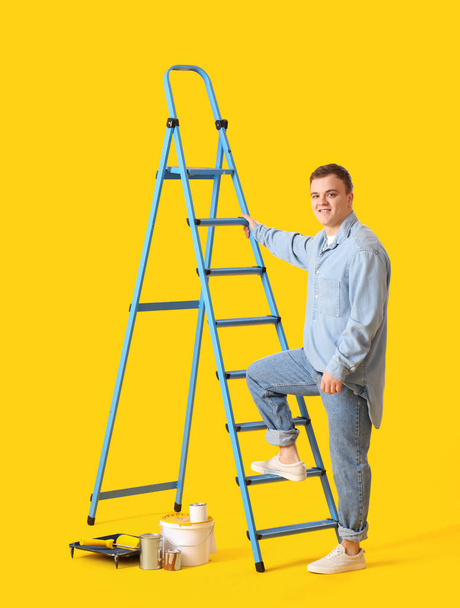 Young man with ladder and cans of paint on yellow background - Zdjęcie, obraz
