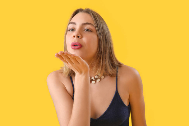 Young woman with necklace blowing kiss on yellow background, closeup - Photo, Image