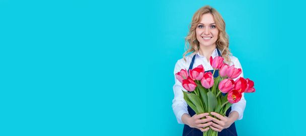 jovem mulher sorrir em avental com flores de tulipa primavera no fundo azul. Retrato facial isolado de mulher, banner com espaço de cópia simulado - Foto, Imagem