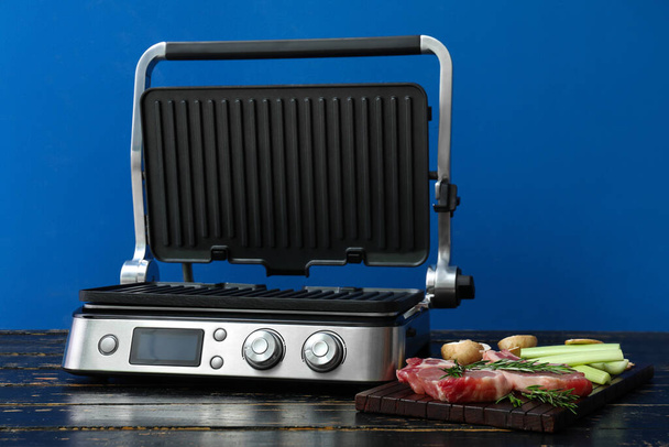 Board with raw steaks near modern electric grill on wooden table against blue background - Φωτογραφία, εικόνα