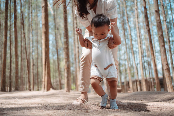Baby macht erste Schritte im Forst auf dem Wiesengras. Mutter unterstützt Kind beim Vorwärtsgehen, Babywachstum und Entwicklungskonzept. - Foto, Bild