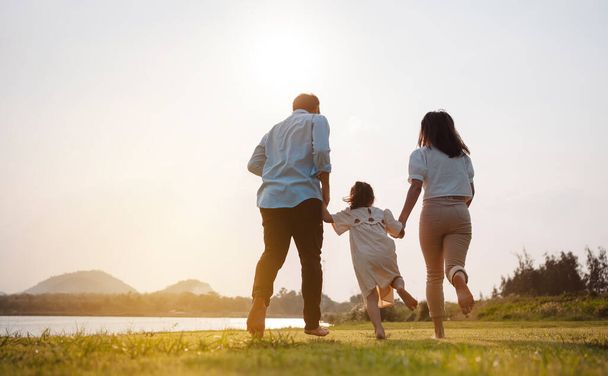 Happy family in the park sunset light. family on weekend running together in the meadow with river Parents hold the child hands.health life insurance plan concept. - Foto, afbeelding