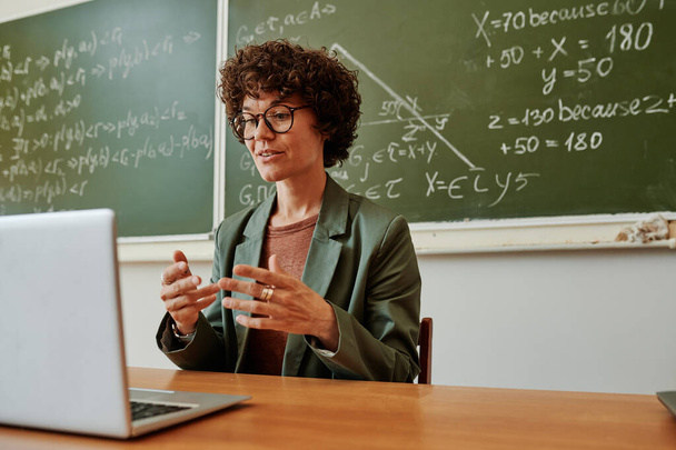 Уверенный молодой учитель, объясняющий новую тему или отвечающий на вопросы онлайн-аудитории, сидя на рабочем месте перед ноутбуком - Фото, изображение