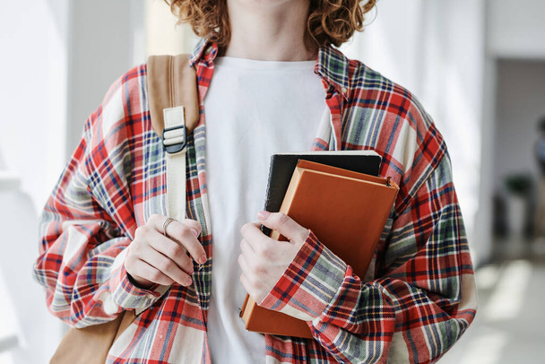 Hands of youthful female student in white t-shirt and checkered shirt holding book and copybook while standing in front of camera - Photo, image