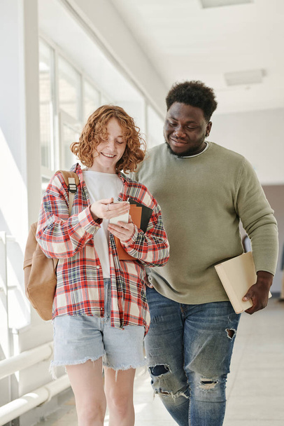 Twee tiener klasgenoten in casualwear bespreken nieuwsgierige dingen in smartphone gehouden door schattig lachend meisje na de lessen - Foto, afbeelding