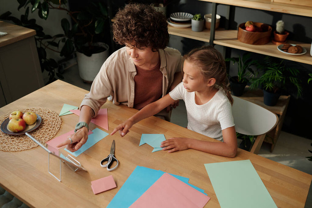 Νεαρή γυναίκα και η κόρη της δείχνει στην οθόνη του tablet κατά τη διάρκεια του online βίντεο για origami τέχνη, ενώ κάθεται δίπλα στο τραπέζι στην κουζίνα - Φωτογραφία, εικόνα
