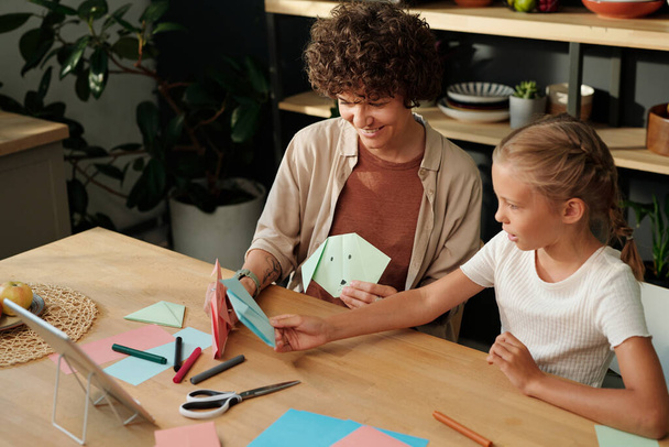 Mladá žena a mladistvé dívka sedí u stolu a porovnává jejich ručně vyrobené origami položky po sledování online masterclass - Fotografie, Obrázek