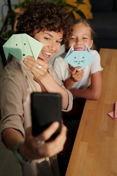 Joven mujer alegre y su linda hija joven con juguetes de origami mirando a la cámara del teléfono inteligente mientras hace selfie por mesa - Foto, Imagen