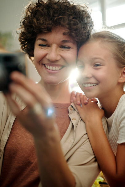 Fröhliche junge brünette Frau mit Retro-Fotokamera, die ein Foto von sich und ihrer niedlichen jugendlichen Tochter macht, die in der Nähe steht - Foto, Bild