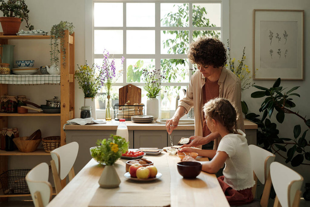 Young woman making sandwiches for youthful daughter and herself for breakfast while standing by kitchen table next to girl eating - Foto, Bild