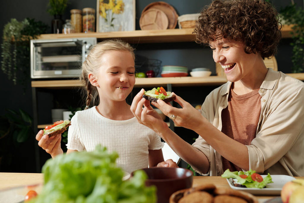 Young laughing brunette woman looking at vegetable sandwich in her hands while sitting by served table next to her youthful daughter - Photo, Image