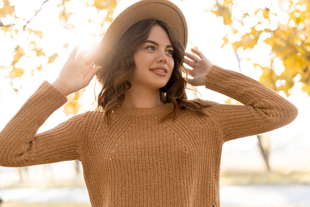 Attractive stylish woman holding hat on head, walking in park, dressed in warm sweater. Young woman with light brown hair smiles enjoying warm weather. Autumn fashion accessories in fashion style. - Фото, зображення