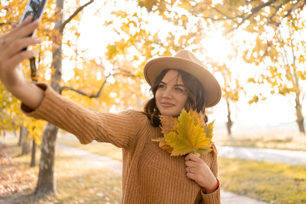 Beautiful young Caucasian woman with fallen leaves taking a selfie with smartphone outdoors in park in autumn. Magnificent brunette girl taking picture of herself while walking. Copy space - Foto, Bild