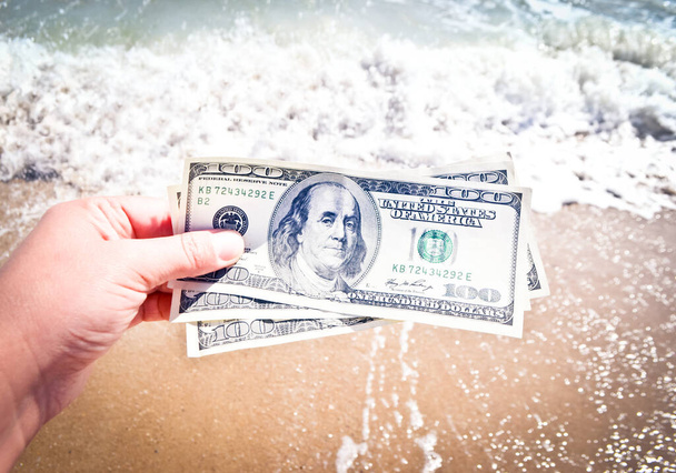 Beyaz köpüklü ve kumlu plajlı deniz dalgalarının arka planında 300 dolarlık banknot tutan bir kız. El dalgasıyla okyanus parası tatili. Mali para seyahati kavramı - Fotoğraf, Görsel