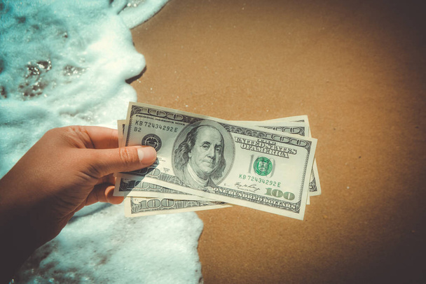 Girl holding money bill of 300 dollars on background of sea ocean waves with white foam and yellow sand wet beach close-up. Hand wave sea ocean money dollars. Concept finance money holiday traveling - Foto, afbeelding