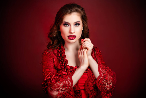 Portrait of a young, attractive vampire woman in a red rococo dress posing isolated against a dark background with red backlights. - Фото, зображення