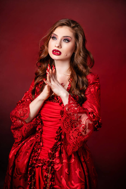 Portrait of a young, attractive vampire woman in a red rococo dress posing isolated against a dark background with red backlights. - Foto, Imagen