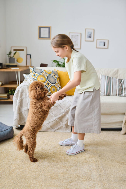 Függőleges kép a kislányról, aki mancsokat tart a kutyájáról és játszik vele otthon a szobában. - Fotó, kép