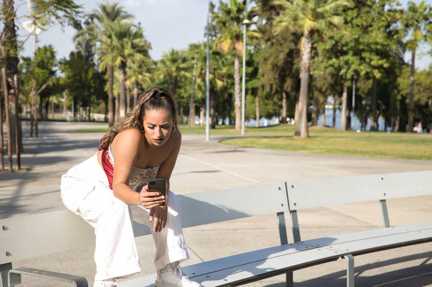 Latin and Hispanic girl, young and beautiful, checking social networks on her cell phone, sitting on a bench. Mobile concept, smartphone, using, consulting, apps. - Photo, Image