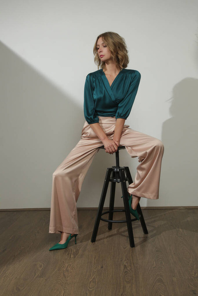 Serie of studio photos of young female model wearing silk satin classic outfit, emerald green wrap blouse and wide legged beige trousers. - Фото, изображение