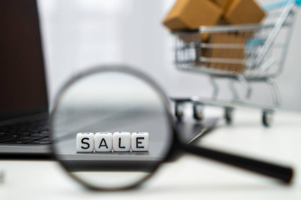 Sale on cubes through a magnifying glass, laptop and toy shopping cart on the background. Online shopping concept.  - Foto, imagen