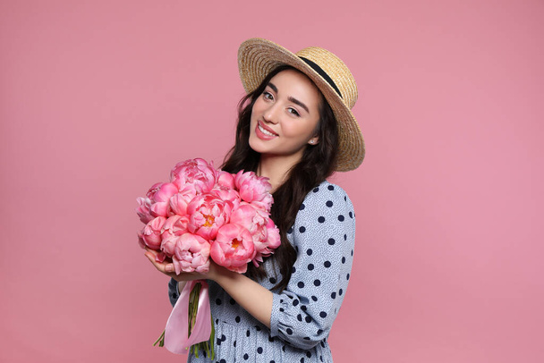 Beautiful young woman in straw hat with bouquet of peonies on pink background - Foto, imagen
