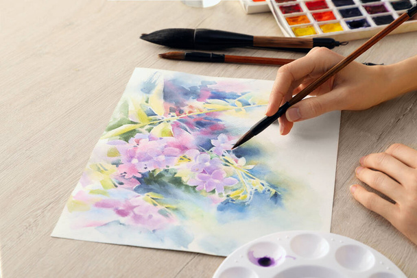 Woman painting flowers with watercolor at white wooden table, closeup. Creative artwork - Foto, Imagen