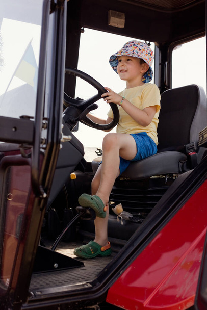 Белый мальчик сидит в салоне трактора на пшеничном поле. Сезон сбора пшеницы - Фото, изображение