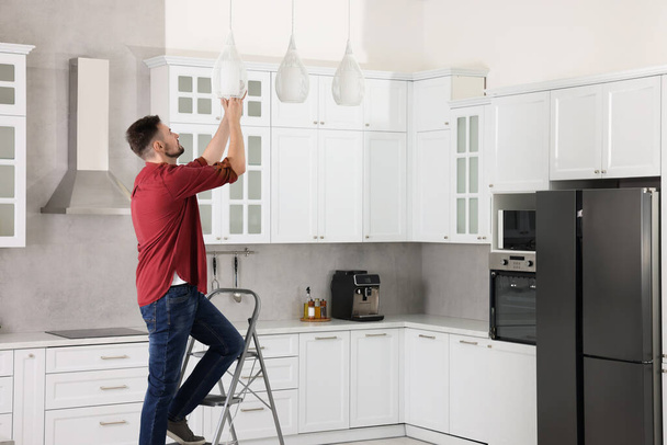 Young man installing ceiling lamp on stepladder in kitchen, space for text - Foto, imagen