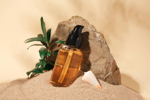 Bottle of serum, stone and green leaves on sand against beige background - Foto, imagen