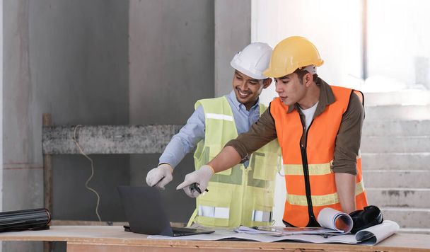 Yelekli ve şapkalı genç adam inşaat alanı inşaatı projesinde dizüstü bilgisayarla çalışıyor. Mühendis iş arkadaşlarıyla konuşuyor.. - Fotoğraf, Görsel