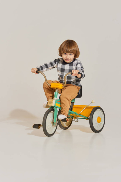 Charming cute boy with long hair riding retro bike and looking at camera over light background. Concept of carefree childhood, holidays, fun, family, retro style - Photo, Image