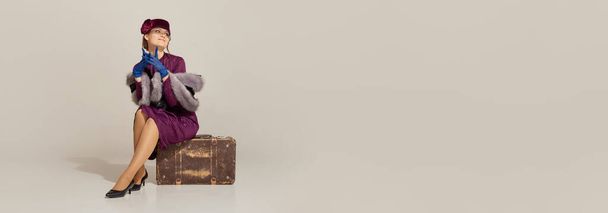 Banner with charming woman wearing old-fashioned clothes and sitting on vintage suitcase over light background. Concept of retro style, fashion, vacation, 60s, 70s, travel - Fotó, kép