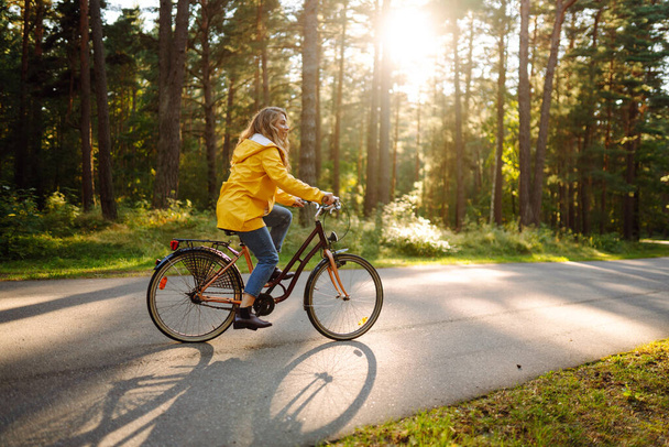 Young happy woman in a yellow coat rides a bicycle in a sunny park. Beautiful woman enjoys autumn nature. Relax, nature concept. Lifestyle. - 写真・画像