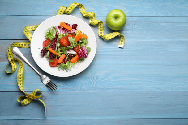 Plate of fresh vegetable salad, apple and measuring tape on light blue wooden table, flat lay with space for text. Healthy diet concept - Fotografie, Obrázek
