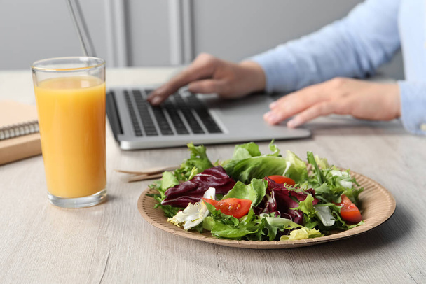 Office employee working with laptop at white wooden table, focus on fresh vegetable salad with glass of juice. Business lunch - Φωτογραφία, εικόνα