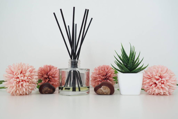Composition of pink artificial flowers, incense sticks in a glass vase with essential oil. Still life on a white table. Aloe flower in a white pot. Room and apartment decor, interior design - Фото, изображение