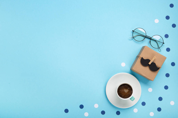 Fathers day holiday card concept with craft gift box, black moustache, cup of coffee and glasses on blue background, top view, copy space. - Photo, Image