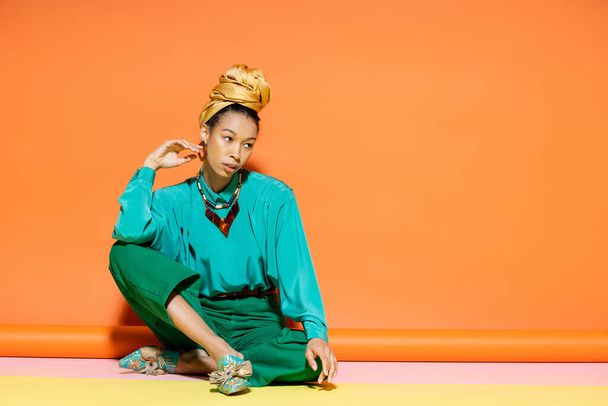 Trendy african american model in bright blouse and pants sitting on colorful background  - Zdjęcie, obraz