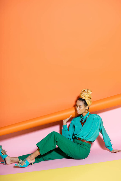 Trendy african american model in bright blouse and headscarf sitting on colorful background  - Фото, изображение