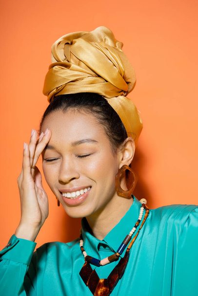 Portrait of smiling african american woman in golden headband touching face on orange background  - Fotografie, Obrázek