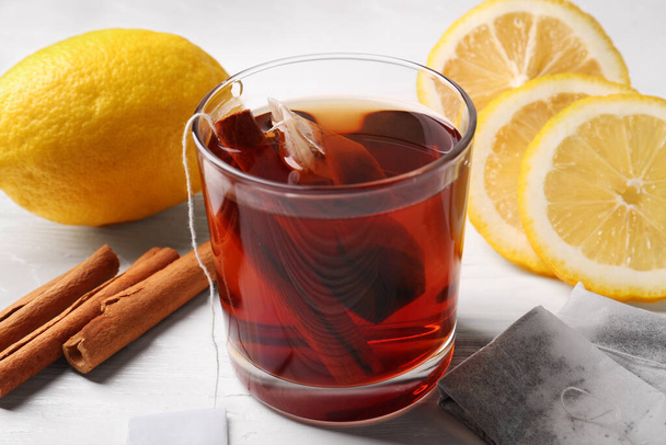 Tea bags, glass of hot drink, cinnamon sticks and lemons on white wooden table, closeup - Фото, изображение