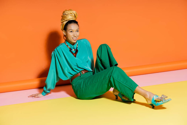 Positive african american woman in summer clothes and shoes sitting on colorful background  - Photo, image